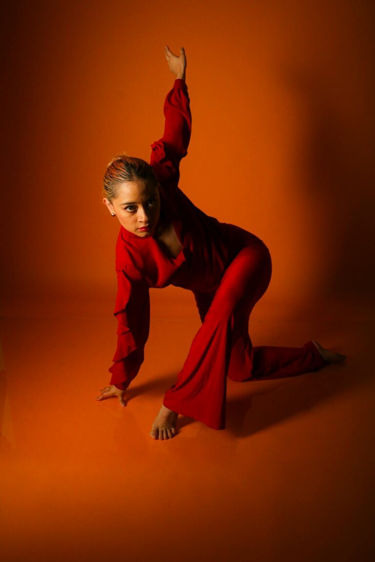 A woman in a red outfit performs a dynamic dance pose in a vibrant orange studio setting.